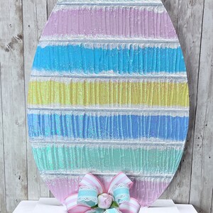 May include: A large, pastel-colored Easter egg with a glittery finish. The egg is decorated with stripes of light blue, yellow, pink, and green. The egg is sitting on a white shelf with a decorative bow.