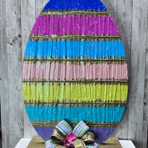 May include: A large, colorful Easter egg decoration with a ruffled texture. The egg is painted in stripes of blue, pink, yellow, and purple with gold accents. A large, gold and plaid bow is tied around the base of the egg.