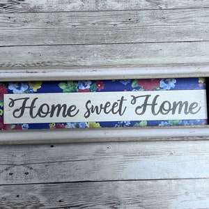 May include: A white framed sign with a blue and floral background. The sign says "Home Sweet Home" in a white cursive font.