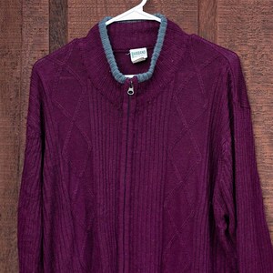 May include: A purple knitted cardigan with a zipper closure and a gray trim. The cardigan is hanging on a white hanger against a wooden background.