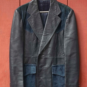 May include: A black leather jacket with suede detailing, suspended on a white hanger against a red wooden backdrop. The jacket has a notched collar, two front pockets, and suede patches on the shoulders and pockets.