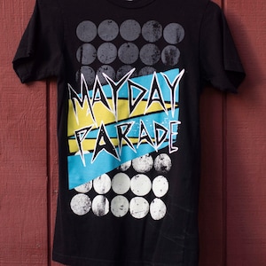 May include: Black t-shirt with the band name "MAYDAY PARADE" in white, yellow, and blue. The design includes rows of gray and white circles. The shirt is hanging on a purple hanger against a red wooden background.