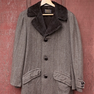 May include: A brown tweed coat with a brown faux fur collar. The coat has a button closure and two flap pockets. The coat is hanging on a wooden hanger against a red wooden background.