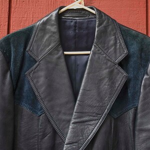 May include: A black leather jacket with dark blue suede shoulder panels and lapels, suspended on a white hanger against a red wooden backdrop. The jacket features a classic design with a notched collar and a tailored fit.