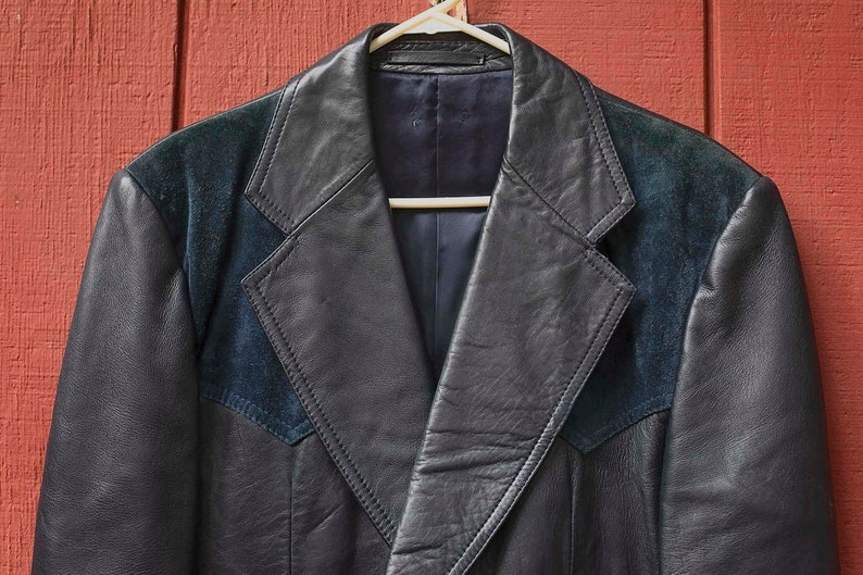 May include: A black leather jacket with dark blue suede shoulder panels and lapels, suspended on a white hanger against a red wooden backdrop. The jacket features a classic design with a notched collar and a tailored fit.