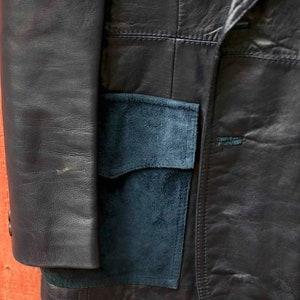 May include: A navy blue leather blazer with a suede pocket is shown in close-up. The jacket features a classic design with visible stitching and a button closure. The suede pocket provides a contrasting texture and colour.