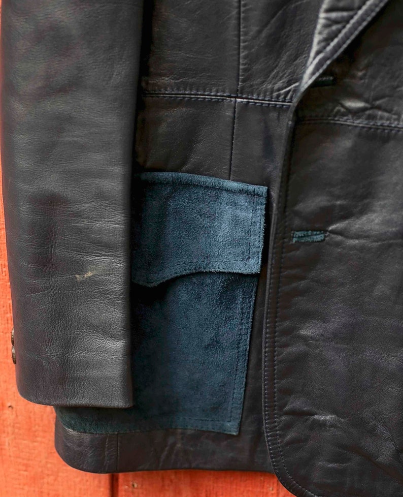 May include: A navy blue leather blazer with a suede pocket is shown in close-up. The jacket features a classic design with visible stitching and a button closure. The suede pocket provides a contrasting texture and colour.