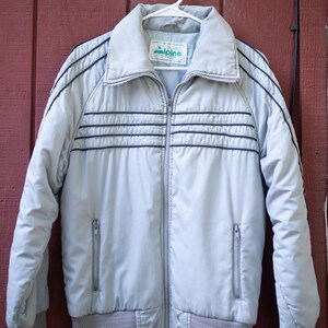 May include: A light gray vintage jacket with black stripes and a zipper closure. The jacket has a label that reads "Alpine" and is hanging on a white hanger.