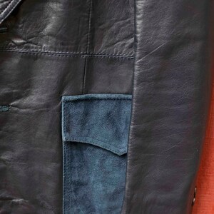 May include: Close-up of a navy blue leather jacket with a teal suede pocket. The jacket has a classic design with visible stitching and a tailored fit, showcasing a blend of textures and colours.