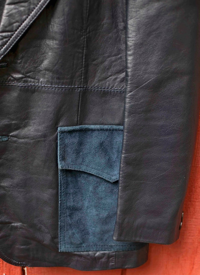 May include: Close-up of a navy blue leather jacket with a teal suede pocket. The jacket has a classic design with visible stitching and a tailored fit, showcasing a blend of textures and colours.