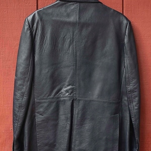 May include: A black leather blazer hangs on a white hanger against a red wooden background. The blazer has a classic design with a notched lapel and a two-button closure. The leather appears to be slightly textured.