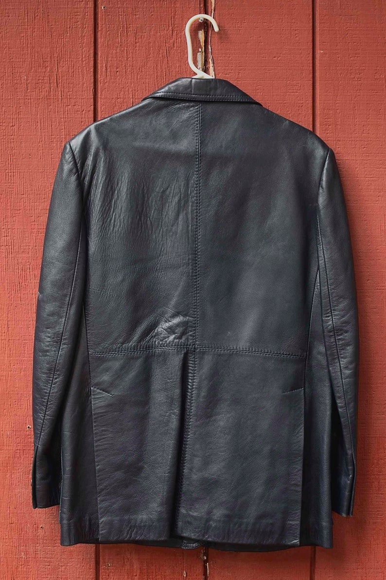 May include: A black leather blazer hangs on a white hanger against a red wooden background. The blazer has a classic design with a notched lapel and a two-button closure. The leather appears to be slightly textured.