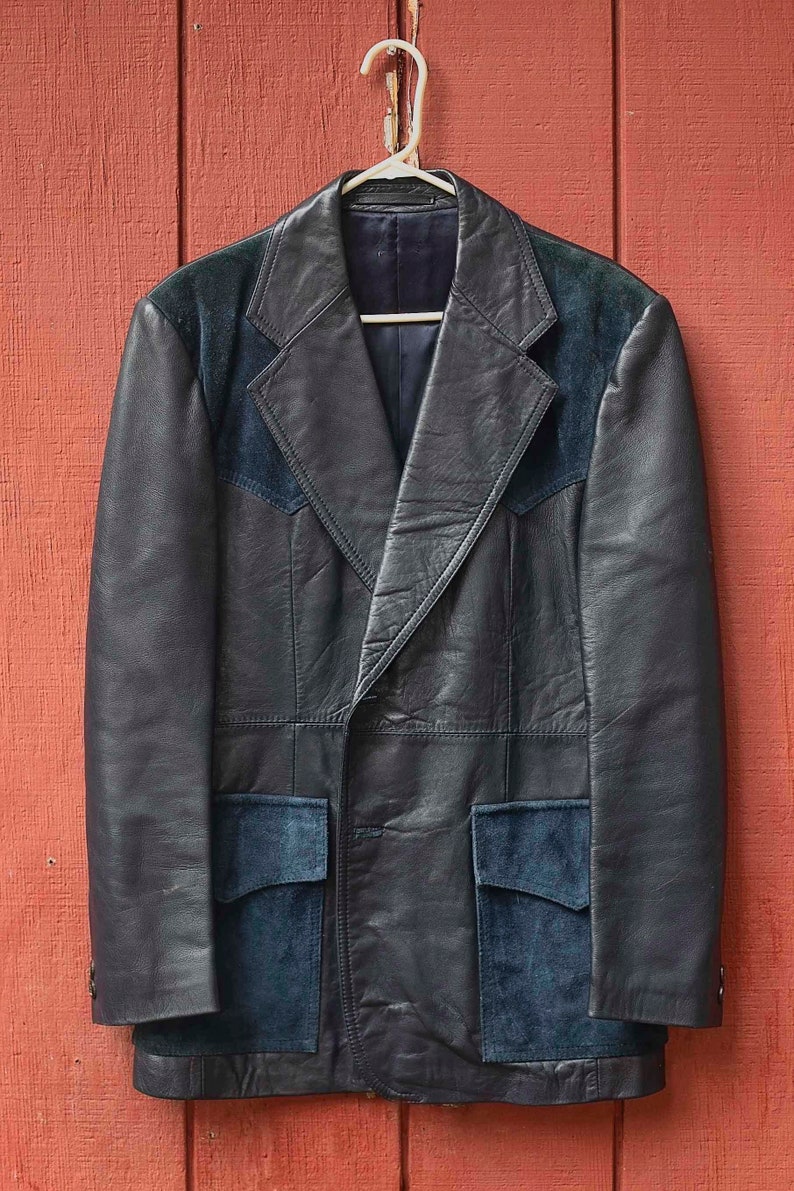 May include: A black leather jacket with suede detailing, suspended on a white hanger against a red wooden backdrop. The jacket has a notched collar, two front pockets, and suede patches on the shoulders and pockets.