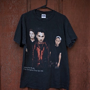 May include: Black vintage t-shirt featuring a band photo of Depeche Mode. The shirt has the text "Depeche Mode The Singles Tour 86>98" printed in white. The shirt is hanging on a black hanger against a brown wooden background.