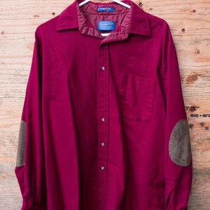 May include: A burgundy long-sleeved button-down shirt with brown leather elbow patches. The shirt has a single chest pocket and a brand label that reads "Pendleton".