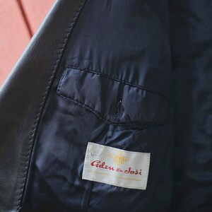 May include: Detail of a black leather jacket's interior. The lining is a dark navy blue, with a small pocket. A label inside the jacket reads "Aden & Josi". The jacket appears to be well-made.