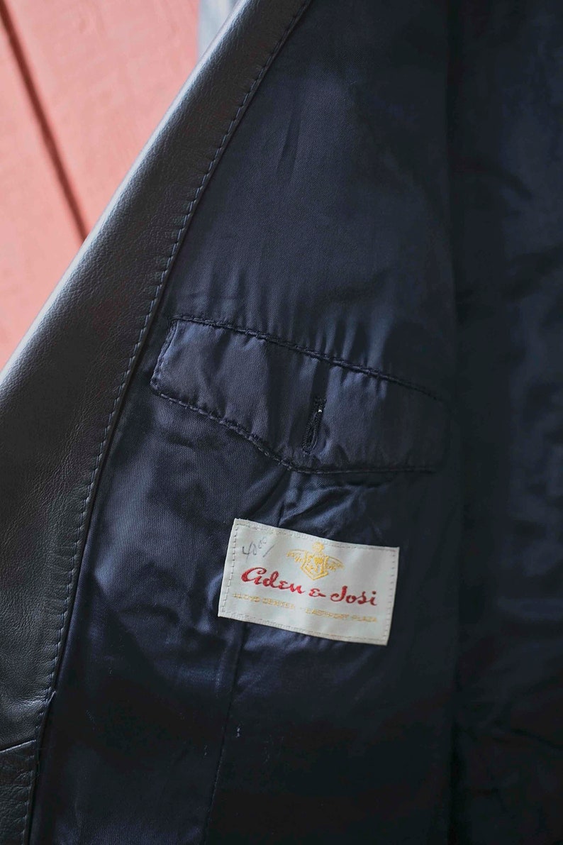 May include: Detail of a black leather jacket's interior. The lining is a dark navy blue, with a small pocket. A label inside the jacket reads "Aden & Josi". The jacket appears to be well-made.