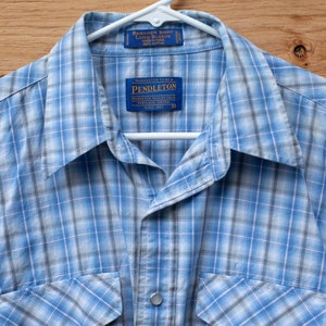 May include: A blue and white plaid long-sleeved shirt with a button-down collar. The shirt has two chest pockets and a Pendleton label on the front.