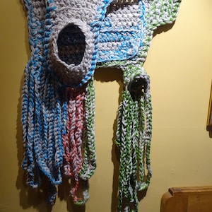 May include: A handmade, gray crocheted wall hanging with blue, green, and red accents. The piece features a central opening and dangling strands of yarn in various colors, creating a unique, textured design.