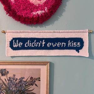 May include: A white crochet banner with the text "We didn't even kiss" in a blue speech bubble. The banner is hung on a gold rod. A pink, textured heart-shaped wall hanging is above the banner.