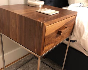 May include: A wooden nightstand with a single drawer and a metal frame. The drawer has a wooden handle and the top of the nightstand is made of walnut wood. The nightstand is sitting on a gray rug.