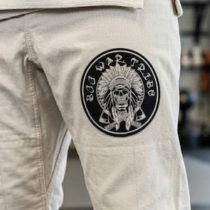 May include: Beige martial arts pants with a large circular patch featuring a skull wearing a Native American headdress, with the words "BAD WAR TRIBE" around the edge. A smaller patch with text is visible on the left leg. The pants are made of a durable fabric.