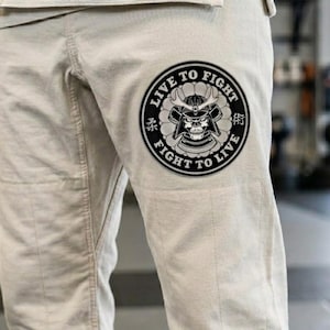 Puede incluir: Pantalones de artes marciales color crema con un parche circular grande con una calavera de samurái y el texto "LIVE TO FIGHT, FIGHT TO LIVE". Los pantalones también tienen un parche más pequeño con texto y un logotipo.