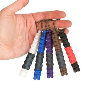 May include: Five keychains featuring braided paracord in white, blue, purple, brown, and black, each attached to a bronze-colored metal ring. Each keychain includes a central black or red textured rubber grip section integrated into the cord.