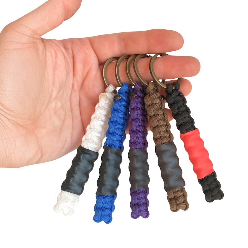 May include: Five keychains featuring braided paracord in white, blue, purple, brown, and black, each attached to a bronze-colored metal ring. Each keychain includes a central black or red textured rubber grip section integrated into the cord.