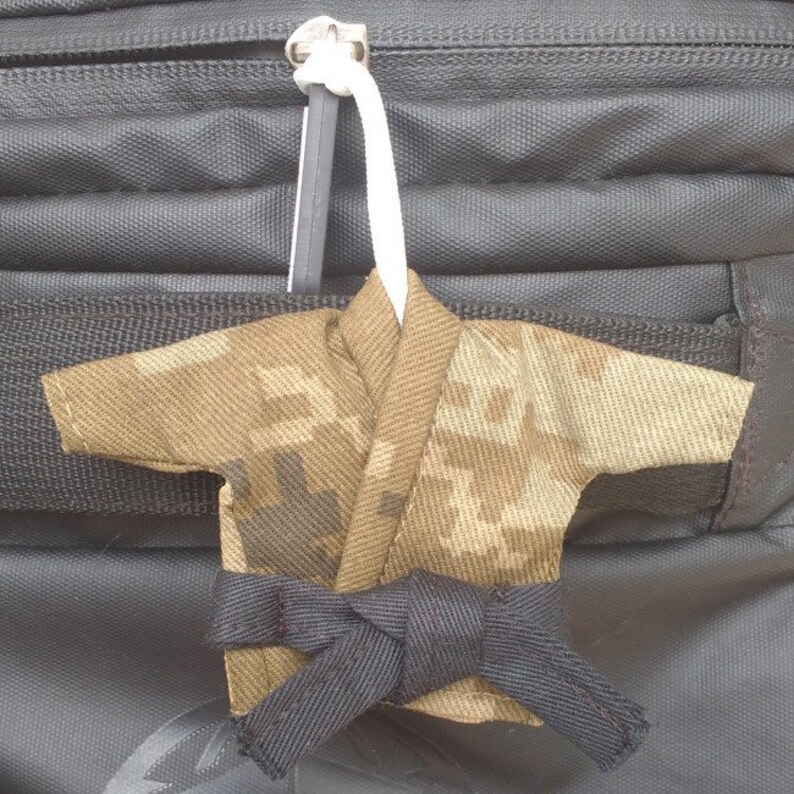 Puede incluir: Uniforme de karate en miniatura con estampado de camuflaje y cintur&oacute;n negro, sujeto a un tirador de cremallera en una bolsa negra.