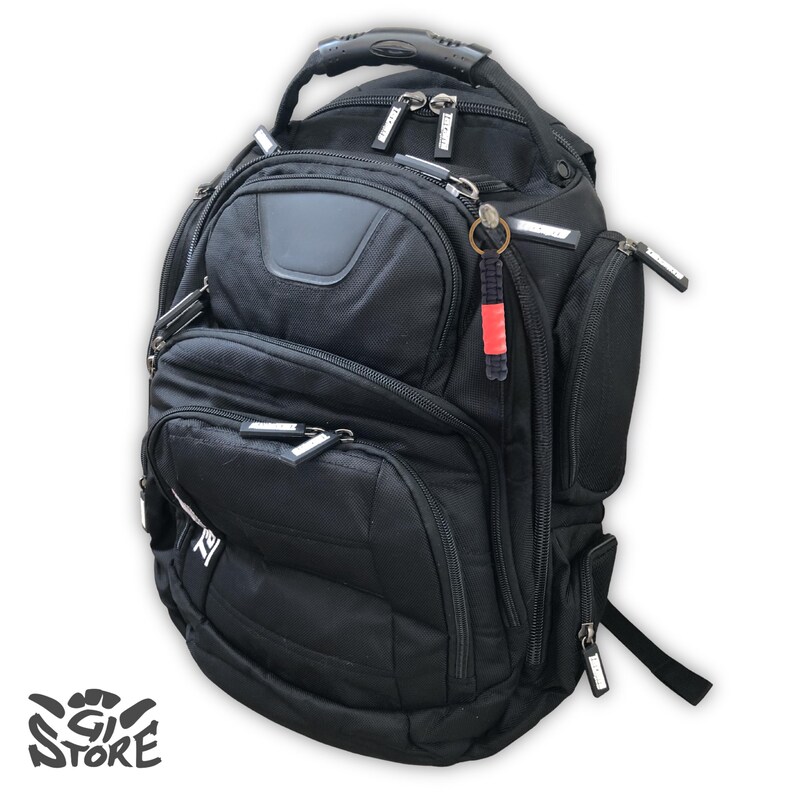 May include: A black backpack featuring multiple zippered compartments, a reinforced top handle, and adjustable shoulder straps. The bag includes a small red and black keychain accessory attached to a zipper pull and the text GI STORE in the corner.