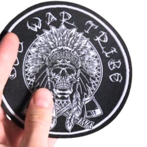 May include: A circular black and white patch with a skull wearing a Native American headdress. The words "WAR TRIBE" are in a circle around the skull. The patch is held by a hand against a white background.
