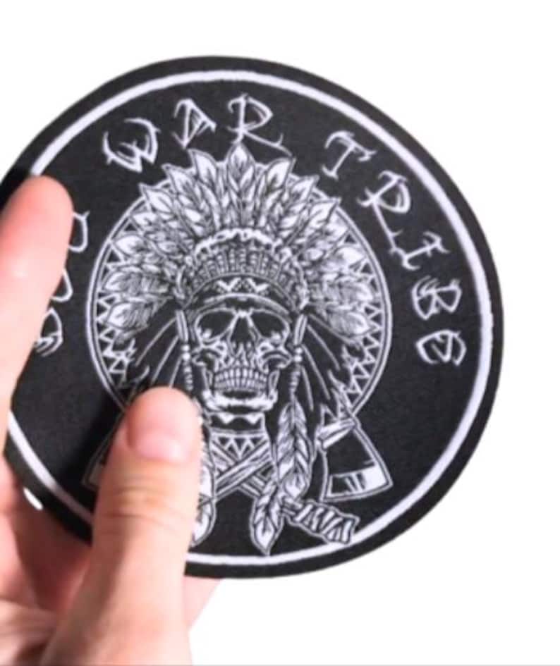 May include: A circular black and white patch with a skull wearing a Native American headdress. The words "WAR TRIBE" are in a circle around the skull. The patch is held by a hand against a white background.