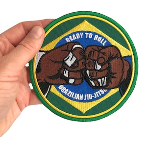 Puede incluir: Un parche bordado circular con un fondo de bandera brasileña estilizada en verde, amarillo y azul. Dos puños marrones se muestran en el centro en un gesto de choque de puños. El texto "READY TO ROLL" y "BRAZILIAN JIU-JITSU" está bordado en blanco.