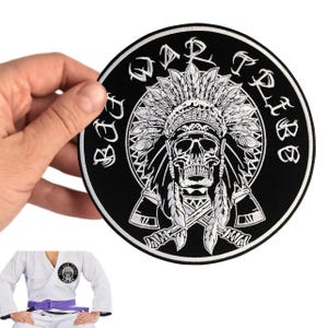 Puede incluir: Un parche bordado circular en blanco y negro que presenta una calavera con un tocado de plumas y dos hachas cruzadas debajo. El texto "BJJ WAR TRIBE" está arqueado en la parte superior. El parche se muestra sostenido y sobre un gi blanco.
