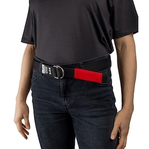Puede incluir: Cinturón negro con hebilla de anilla en D plateada y un acento rojo. El cinturón se lleva con vaqueros de lavado oscuro y una camiseta negra. El cinturón tiene un parche rojo rectangular y una etiqueta blanca.