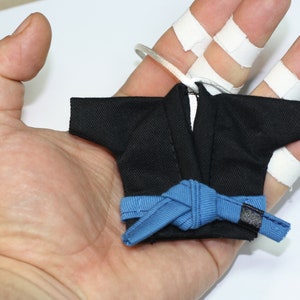 Op de afbeelding: Een miniatuur zwart karatepak met een blauwe band die in een knoop is geknoopt. Het pak is klein genoeg om op een vinger te passen.