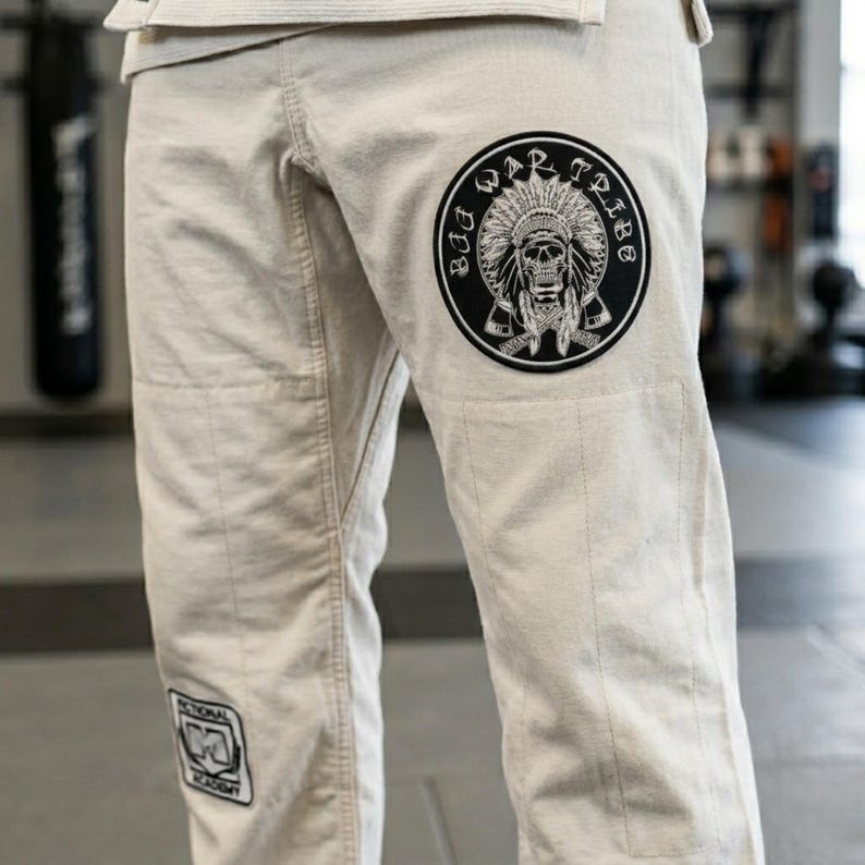 May include: Beige martial arts pants with a large circular patch featuring a skull wearing a Native American headdress, with the words "BAD WAR TRIBE" around the edge. A smaller patch with text is visible on the left leg. The pants are made of a durable fabric.