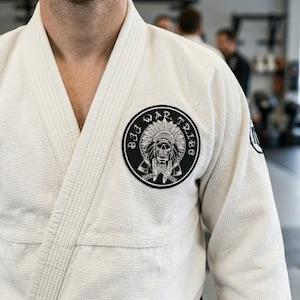 May include: A cream-coloured martial arts uniform with a black and white circular patch. The patch features a skull wearing a Native American headdress, with the words "Bad War Tribe" around the edge.