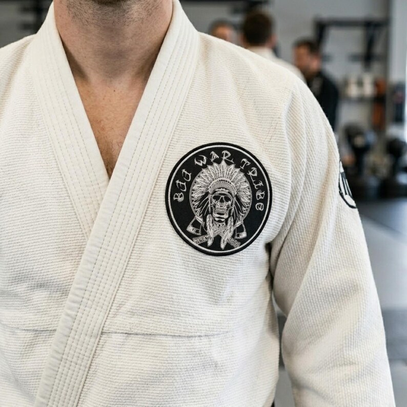 May include: A cream-coloured martial arts uniform with a black and white circular patch. The patch features a skull wearing a Native American headdress, with the words "Bad War Tribe" around the edge.