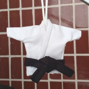 Puede incluir: Uniforme de karate blanco con un cintur&oacute;n negro atado alrededor de la cintura. El uniforme est&aacute; colgado de una cuerda.