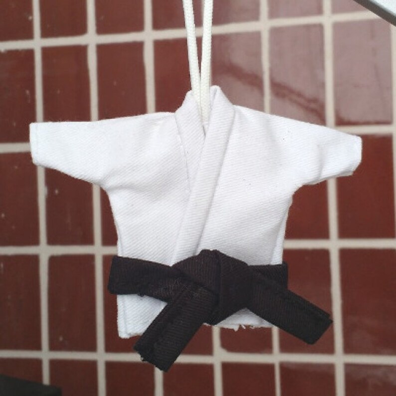 Puede incluir: Uniforme de karate blanco con un cintur&oacute;n negro atado alrededor de la cintura. El uniforme est&aacute; colgado de una cuerda.