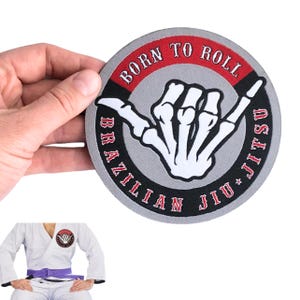 Könnte beinhalten: Ein runder bestickter Aufnäher mit einer weißen Skeletthand, die ein Shaka-Zeichen auf grauem Hintergrund zeigt. Der rot-schwarze Rand enthält den Text BORN TO ROLL und BRAZILIAN JIU JITSU. Der Aufnäher ist auch auf einem weißen Gi zu sehen.
