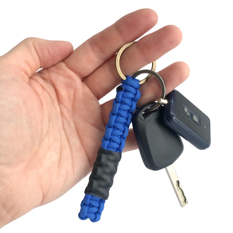 May include: A blue paracord keychain featuring a braided design with a smooth black cylindrical center section. The keychain is attached to a gold-toned metal split ring, which also holds two black plastic car key fobs with metal keys.