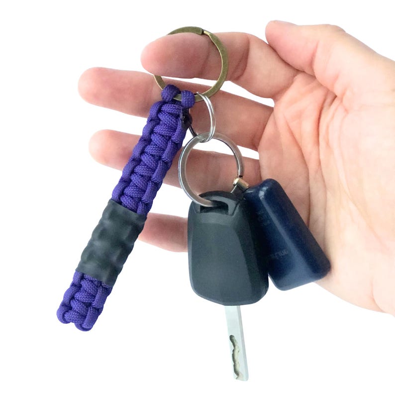May include: A purple paracord keychain featuring a braided design with a solid black rubberized grip section in the center. The keychain is attached to a circular metal ring along with a black car key fob and a dark blue plastic key fob.