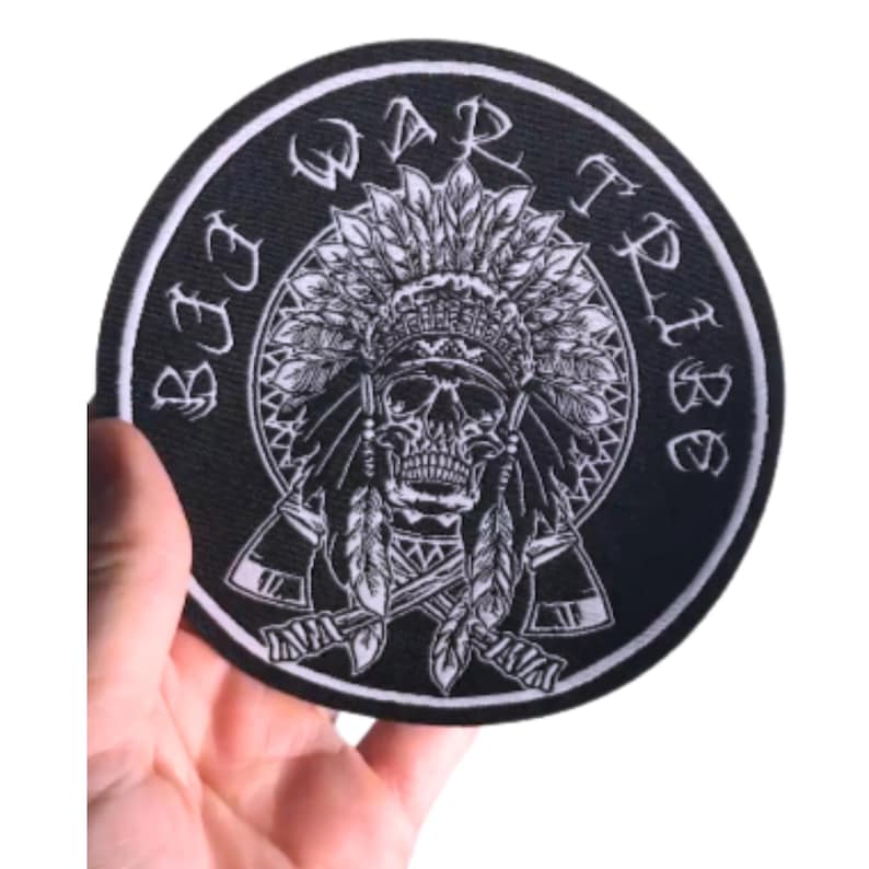 May include: A circular black patch featuring a white skull wearing a feathered headdress, with the words "WAR TRIBE" and "BEE TRIBE" in white around the edge. The skull has detailed features and is surrounded by feathers and axes.