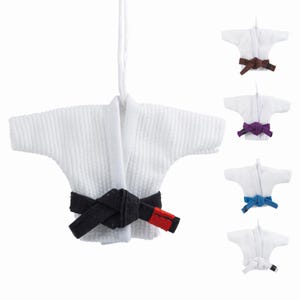 May include: A white miniature martial arts uniform with a black belt and red detail. Other uniforms with brown, purple, blue, and white belts are also visible. The uniform is suspended by a white string.