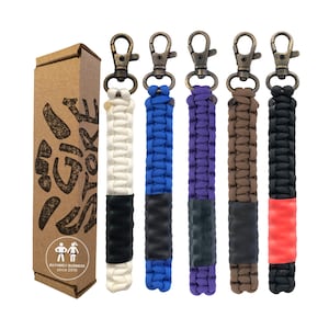 May include: A collection of keychains with bronze-colored clasps and braided cords in various colors: white, blue, purple, brown, and black. Each keychain has a black or red accent. A brown cardboard box with text and a logo is on the left.