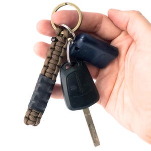 May include: A keychain featuring a brown paracord fob with a braided pattern and a smooth black cylindrical section, attached to a circular metal ring along with a black plastic car key fob and a dark blue rectangular plastic key cover.