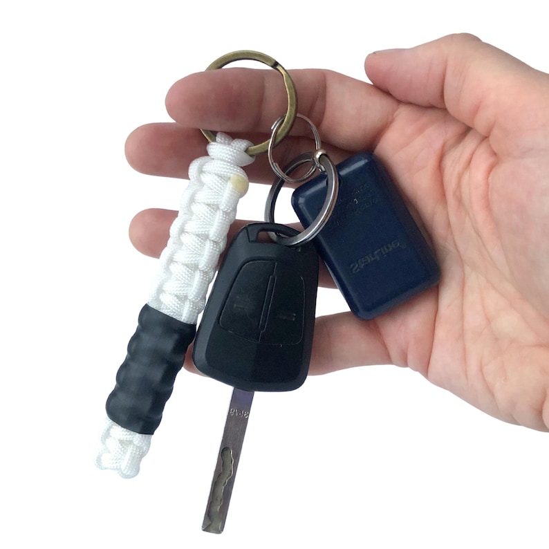 May include: A keychain featuring a white paracord fob with a black rubber grip section, attached to a brass-toned metal ring. The ring also holds a black car key with a metal blade and a small, rectangular dark blue plastic electronic fob.